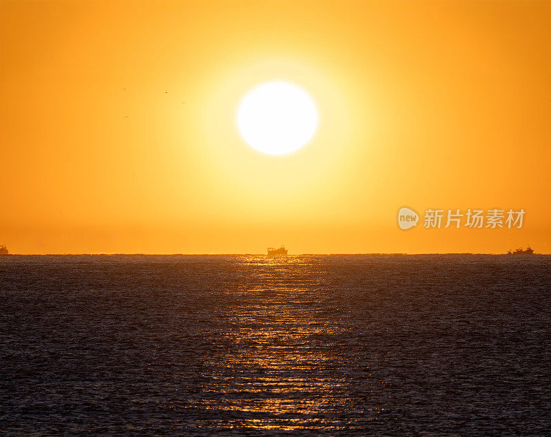 明亮的黄色黎明的太阳从地中海的地平线升起，照亮了一艘渔船在平静的阳光下航行，在橙色的天空下捕虾。