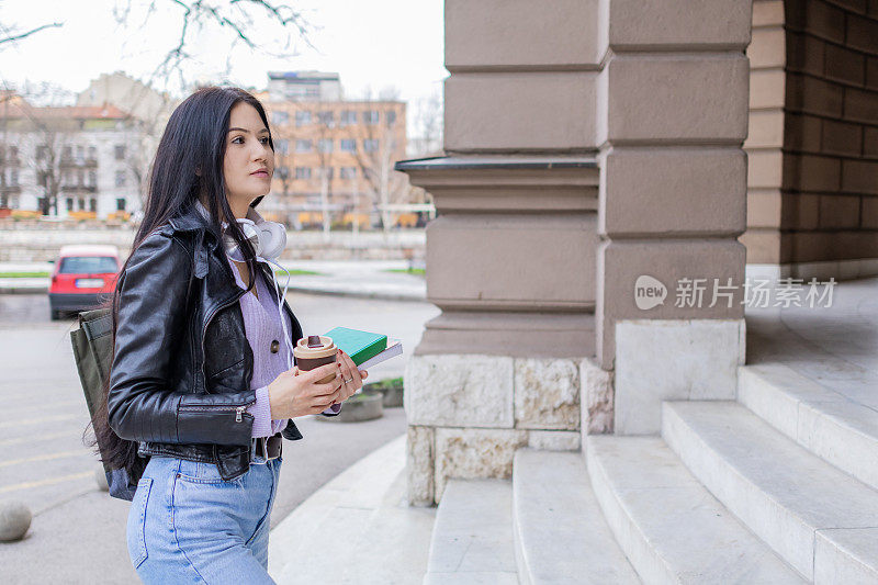 年轻漂亮的女大学生背着双肩包站在大学的楼梯上。她戴着耳机，穿着皮夹克，等待着大学课程的开始。