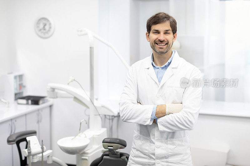 牙科中心。男性牙科医生在工作场所的肖像，口腔医生在现代诊所内部双臂交叉站立