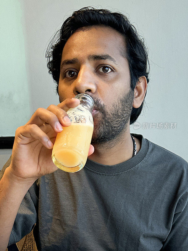 印度男子从玻璃瓶里啜饮橙汁的特写照片，坐在室内布景中，聚焦于前景
