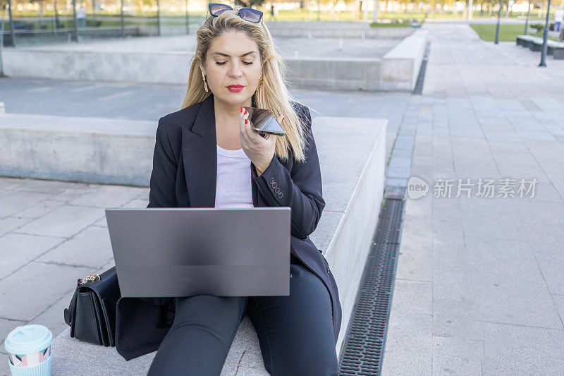 穿着黑色西装的女商人，坐在公司大楼前，一边用笔记本电脑工作，一边用手机打电话。在户外灵活工作。现代虚拟理财咨询。