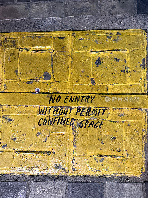 黄色井盖的特写图像，上面写着拼写错误的“未经许可不得进入密闭空间”的标志，铺砌的石头人行道，检查入口，高架视图