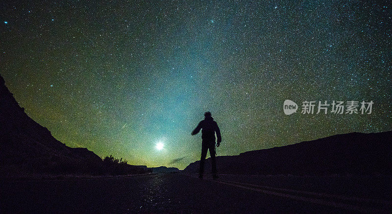 这个小个子男人站在无尽的宇宙前面。在犹他州拱门国家公园的山上，一个人的剪影在星空的映衬下走向月亮