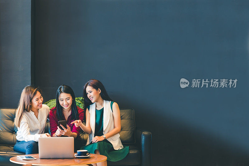 三个美丽的亚洲女孩使用智能手机和笔记本电脑，在咖啡馆的沙发上聊天与复印空间，现代生活方式与电子产品技术或职业女性在休闲的商业理念