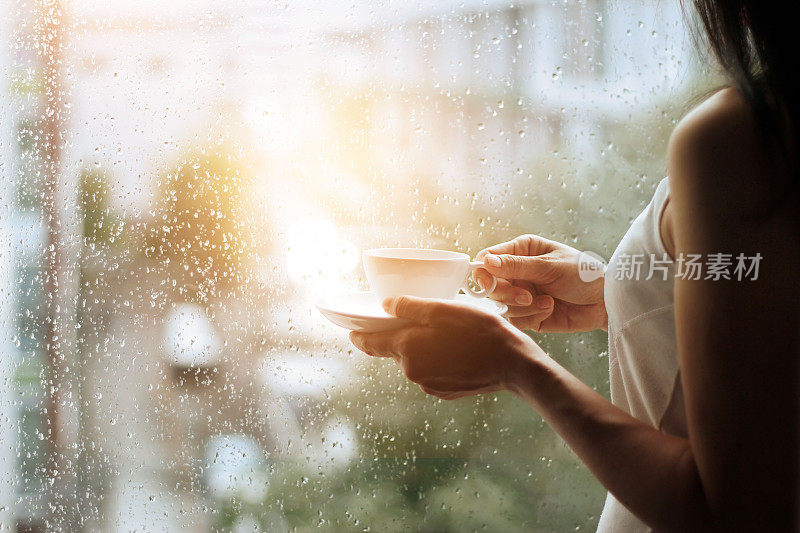 手里拿着咖啡的女人透过玻璃窗看着城市背景上的雨滴