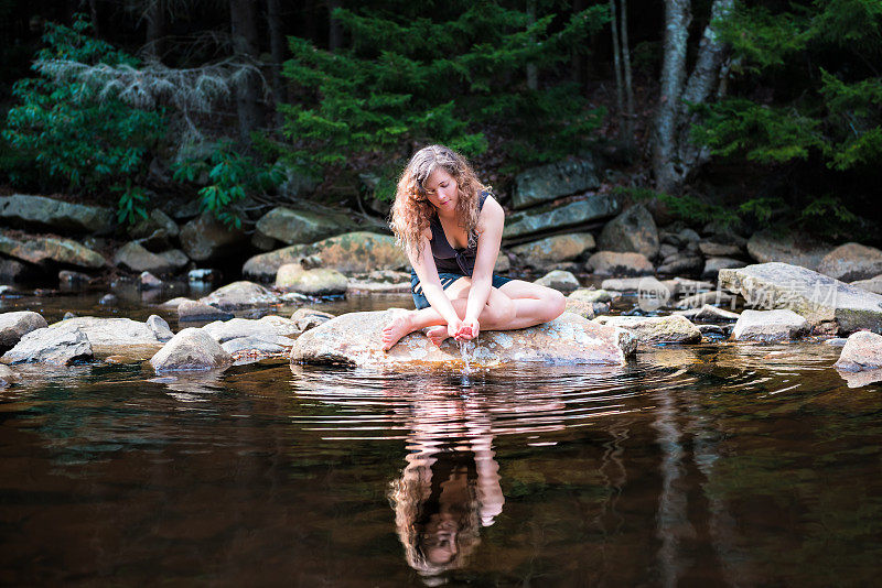 在阳光明媚的日子里，西维吉尼亚州多莉草皮上的红溪河上，年轻女子享受着大自然的宁静，双手浸在水里喝新鲜的水，溅起水花