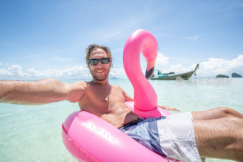 在泰国岛屿清澈的海水中，一个年轻人在田园般的海滩上与粉红色的充气火烈鸟自拍。人们旅行奢侈，乐趣和酷的态度概念