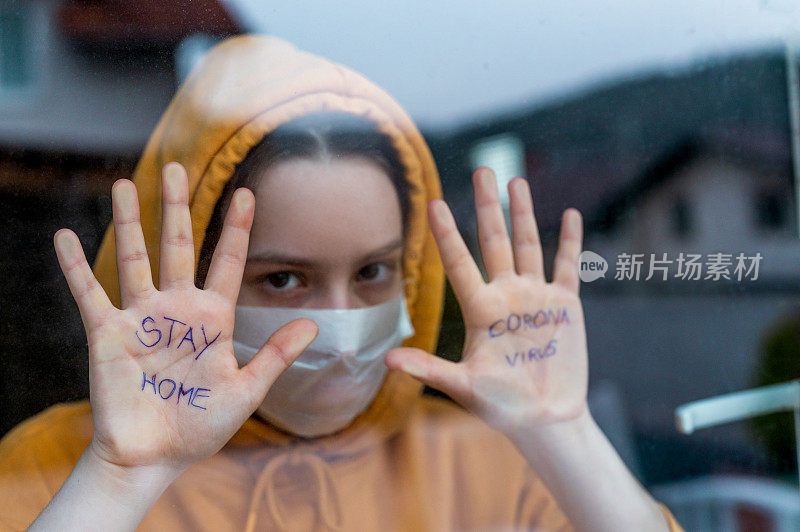 一个十几岁的女孩站在窗前，手上写着“待在家里”和“冠状病毒”