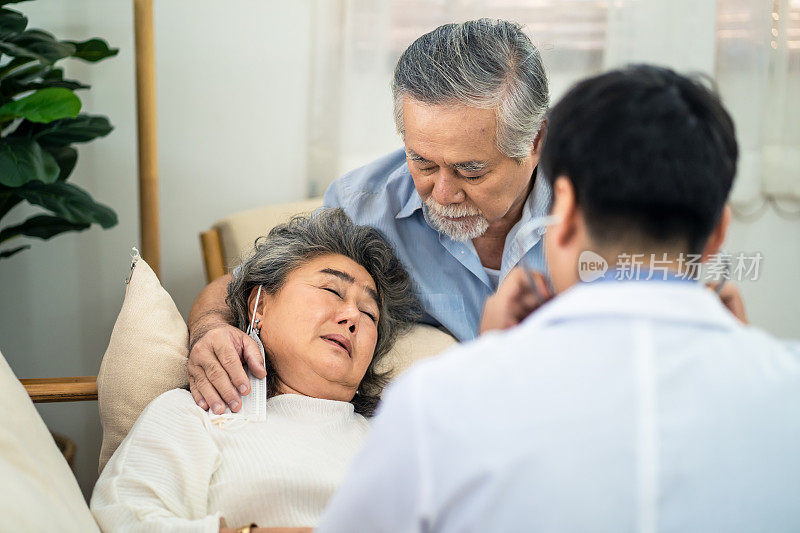 亚洲老年妇女与不好的条件感到生病躺在沙发上有医疗检查由医生来访的家。年长的男人对丈夫关怀备至，担心妻子生病。