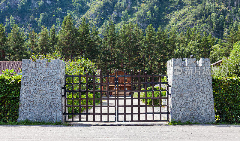 程式化的休闲中心入口，通过城堡的大门，有石柱和铁栏，绿树和山下的木屋。装饰花园。