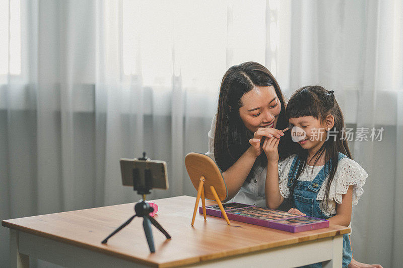 快乐年轻的亚洲母亲和她的女儿录制视频博客在家里与化妆主题