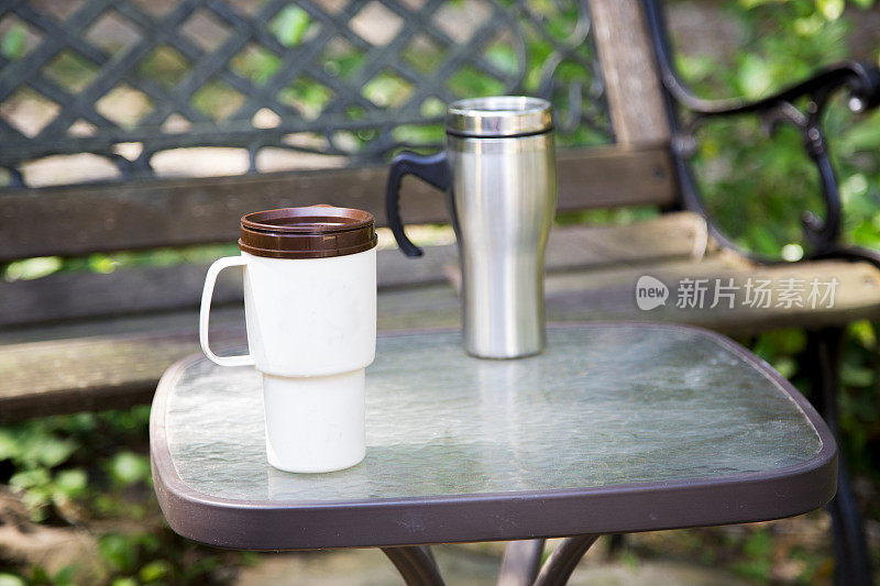 公园长椅前的桌子上放着两个可重复使用的咖啡杯。