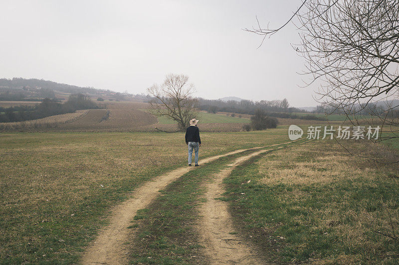 英俊的成年农民戴着牛仔帽走在穿过农田的土路上