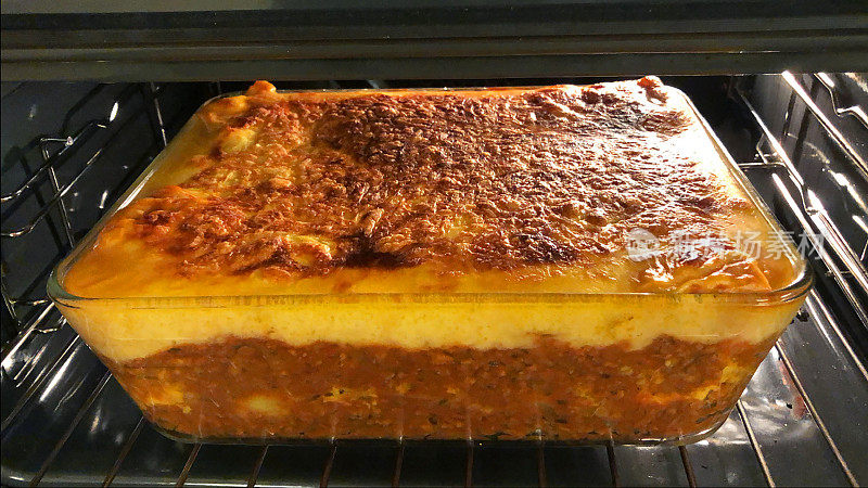 烤箱架上的玻璃防烤盘的图片，里面有羊肉和土豆茄饼，上面浇上酥脆的金黄色奶酪酱，烤箱很热
