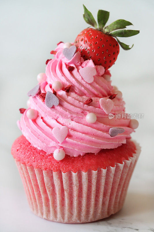个人自制的粉色天鹅绒草莓纸杯蛋糕的特写，草莓上的粉色奶油糖霜，糖工艺心形和球形点缀，大理石蛋糕架，关注前景