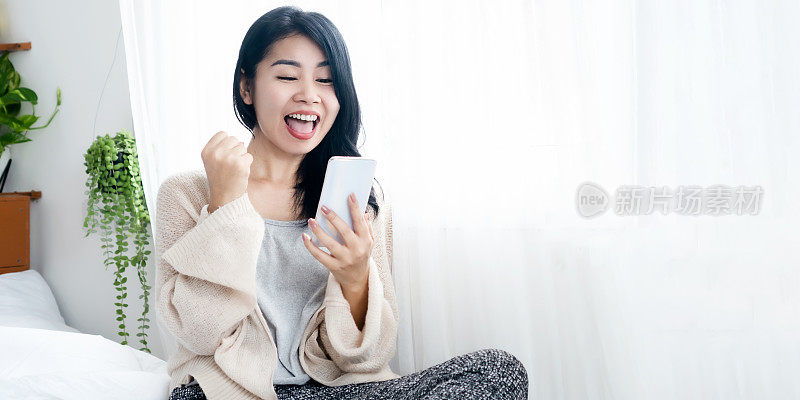 开朗的亚洲女人兴奋地得到好消息，手握手机在床上笑
