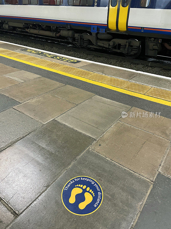 火车站站台上深蓝色和黄色圆形贴纸的图像，新冠病毒社交距离规定，保持2米距离，Covid-19安全，等待列车，“注意台阶”标识，站台边缘的触觉铺设，聚焦前景