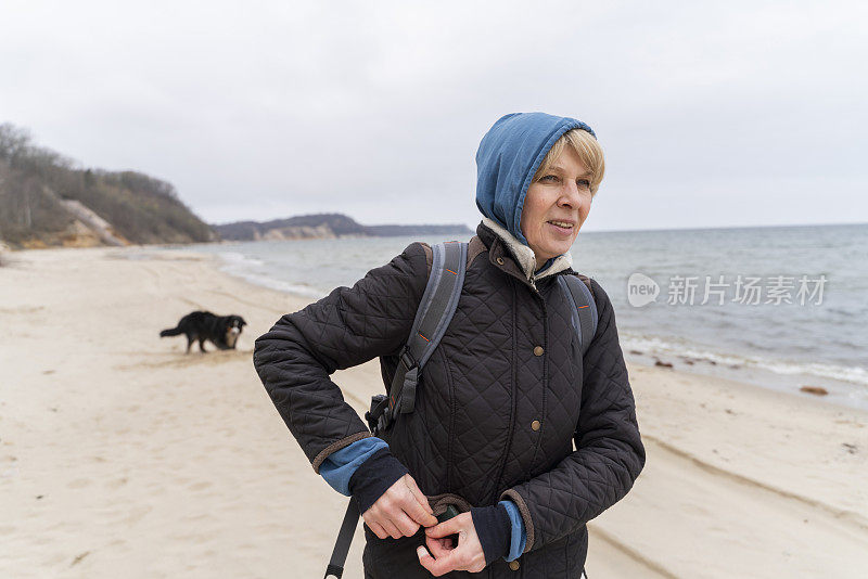 凉爽的春日，在海边遛狗。一个成熟的金发女人穿着黑色的夹克，带着蓝色的兜帽，在前景中，在腰部，黑色的狗在散焦的远处。