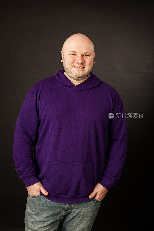工作室肖像快乐超重中年成年白人男子剃了头，穿着紫色连帽衬衫，反对黑色背景