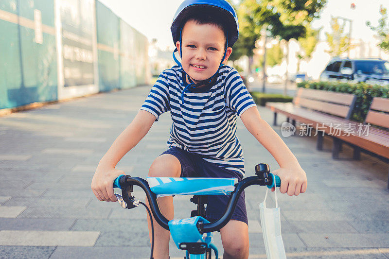 在新冠肺炎疫情期间，可爱的小男孩戴着防护口罩和安全帽在阳光明媚的夏日在公园骑自行车，新常态的生活方式概念。