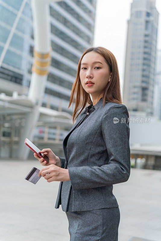 一名年轻的亚洲女商人穿着灰色的办公套装，在商业区用她的智能手机使用信用卡进行在线交易，她微笑着自信