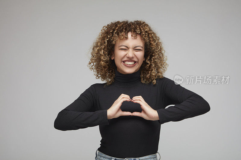 年轻的卷发女子闭着眼睛，高兴地用手做心形手势。