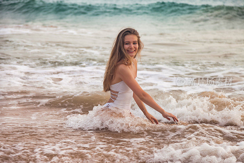 漂亮的年轻苗条的女人在白色泳衣摆姿势在海水波浪背景，看着相机。轻松愉快的热带假期。旅游度假度假概念。复制广告文字空间