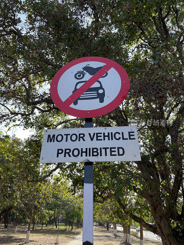 特写图像:红框圈，白色背景，摩托车和汽车的标志被红色对角线穿过，黑白条纹杆，白色矩形“机动车禁止”警告标志，林荫人行道，重点在前景
