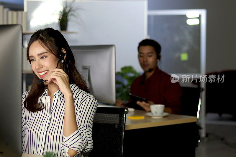 戴着耳机的美丽亚洲女性在呼叫中心客户服务柜台上夜班，看着台式电脑，回答问题，女性用免提电话加班努力工作。