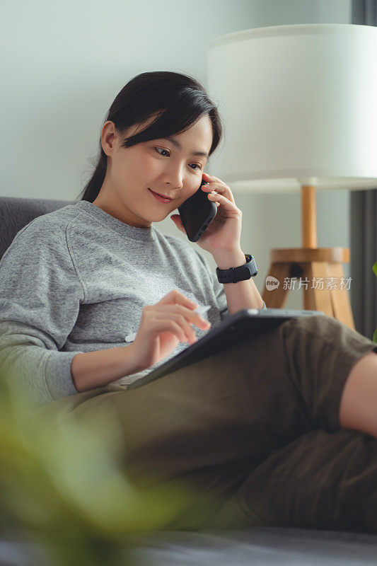 亚洲女性坐在家里的客厅里，用智能铅笔在数字平板电脑上画画，打电话。