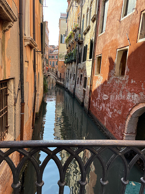 从意大利威尼斯狭窄运河上的桥梁上观看的特写图像，桥梁横跨建筑物排列的水道，多云的蓝色晴朗的天空，缩小的视角，聚焦于前景