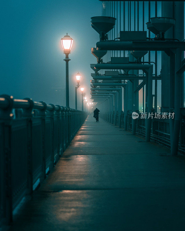 一名男子在雾蒙蒙的夜晚过桥，前景是灯柱