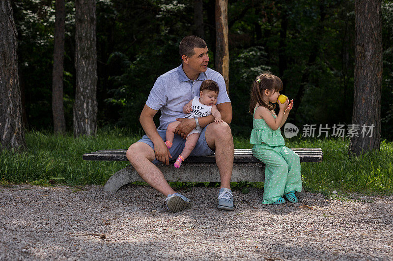 父亲抱着婴儿，大女儿在森林里吃苹果。