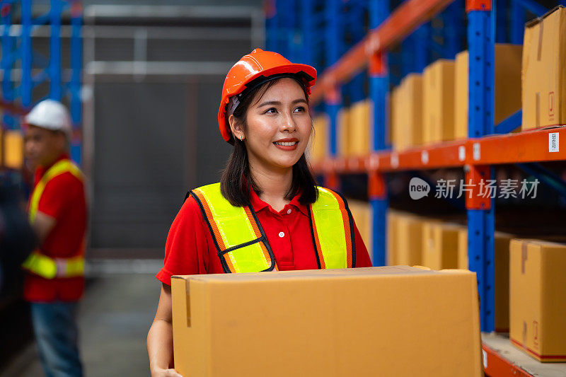 亚洲女仓工，身穿制服，头戴安全安全帽，在仓厂搬运纸箱。批发商品。货架、托盘和箱子