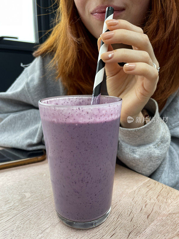 特写照片中，难以辨认的红发女子坐在餐厅的餐桌旁，喝着一杯蓝莓冰沙饮料，通过条纹状的纸吸管啜饮，聚焦于前景