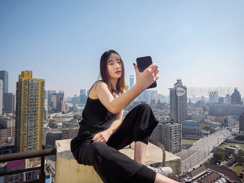 美丽的年轻女子坐在大厦屋顶上，以模糊的上海外滩地标建筑为背景自拍。情感、人、美、生活理念。