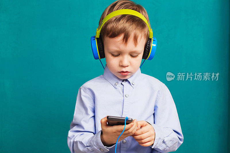 帅气的小男孩戴着明亮的耳机站着，通过智能手机听着音乐，靠在绿色的蓝色墙壁上