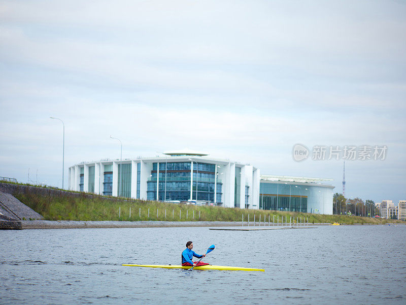 城市划独木舟。在现代建筑和阴天的背景下，年轻的男运动员划着黄色的独木舟顺流而下