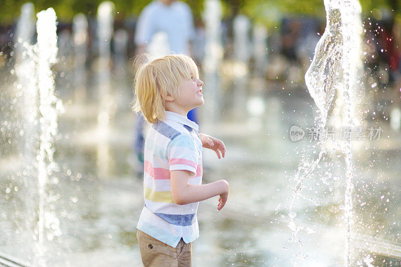 在阳光明媚的夏日，小男孩在广场喷泉的喷水口之间玩耍。活跃的夏季休闲城市的孩子。