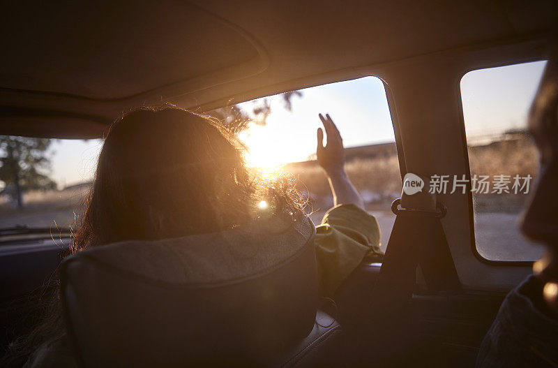 年轻女子举起手臂，在车窗外感受风在日落