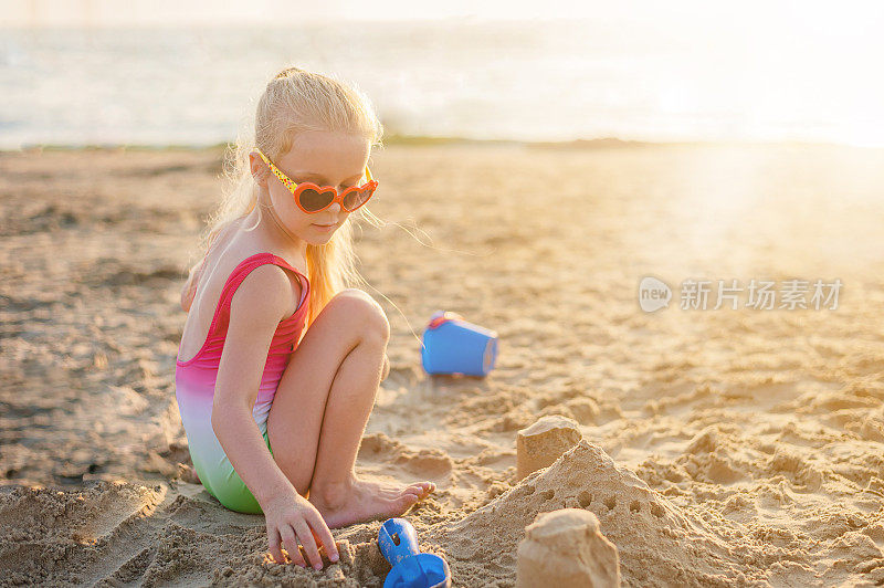 在夏天的黄金时间，傍晚日落时，金发小女孩在海边建了一座沙堡