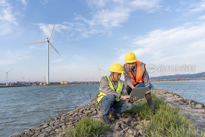 两名亚洲风力发电工程师蹲在海边的风力发电站里使用电脑