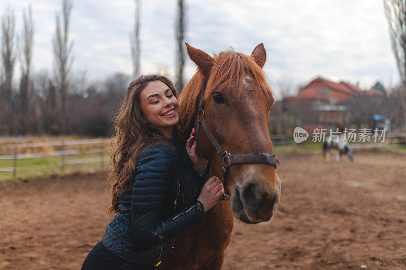 在农场上，一名年轻的白人妇女穿着皮夹克，拥抱着她的棕色马