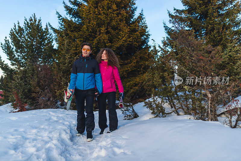 一对美丽的年轻夫妇在雪山上散步，手牵着手，玩着滑雪板