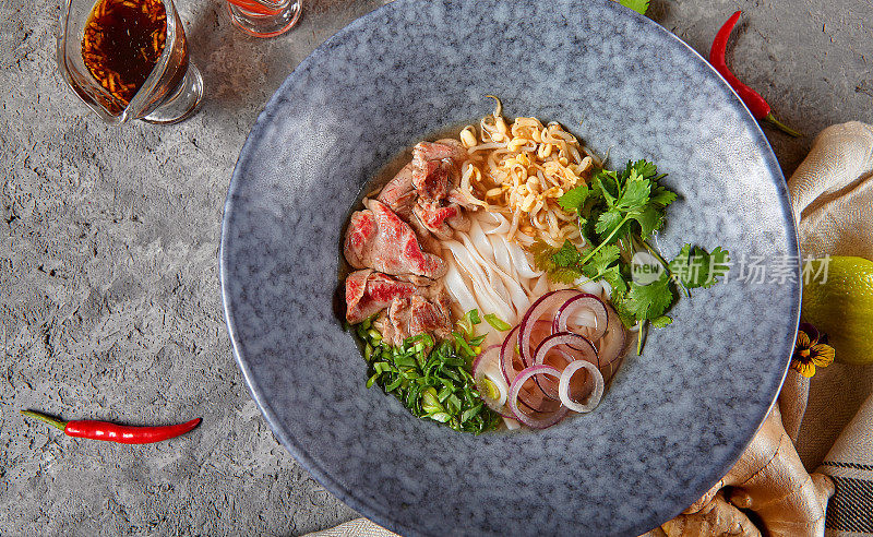 越南菜-牛肉米粉面汤或米粉汤。与新鲜的绿色蔬菜在蓝色碗上纹理深灰色的桌子。麻辣肉汤。前视图。