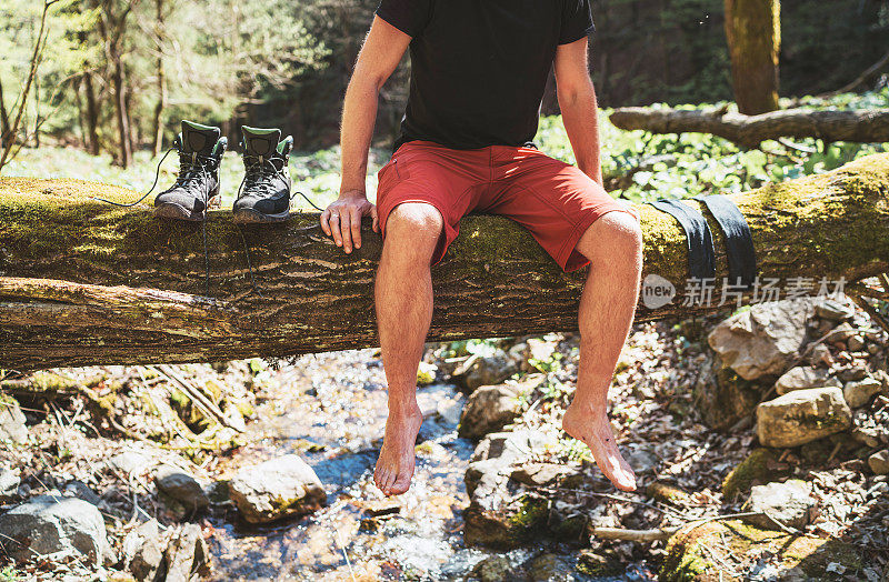 一名男子坐在山林溪流上倒下的圆木上，他在等待袜子、衣服晾干和徒步旅行的靴子。积极的人旅行，徒步或徒步概念形象。