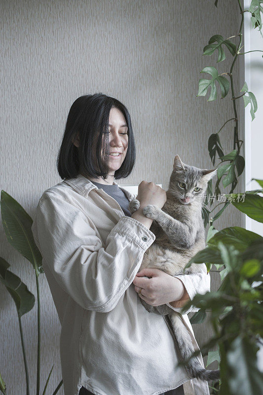 一位年轻的妇女抱着一只黄眼睛的灰猫。猫和她的女主人在家里。照顾宠物。