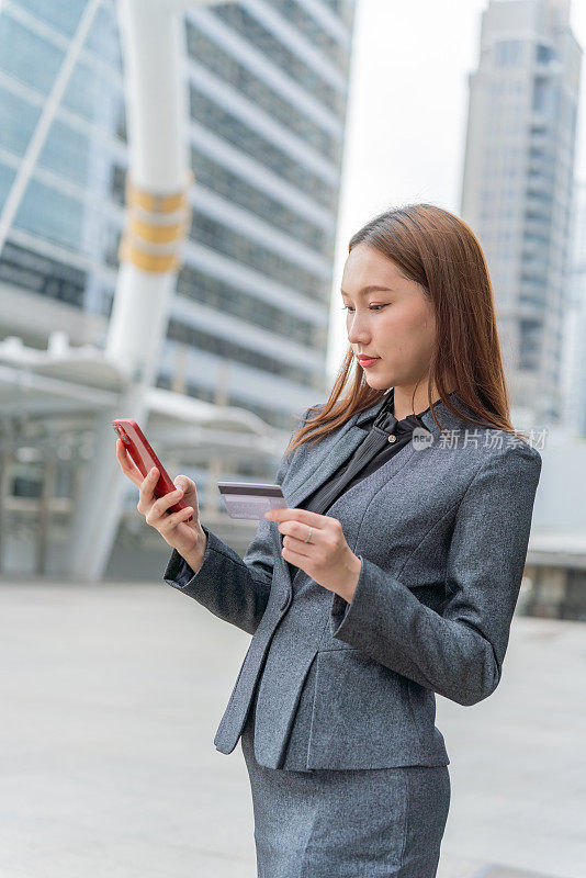 一名年轻的亚洲女商人穿着灰色的办公套装，在商业区用她的智能手机使用信用卡进行在线交易，她微笑着自信