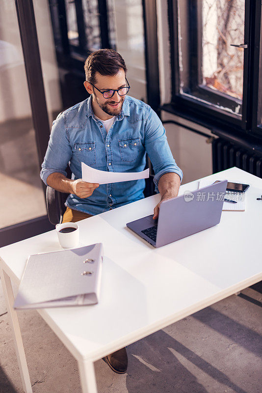 一个戴着眼镜的满意的年轻人坐在办公桌前，在他的工作场所做文书工作。