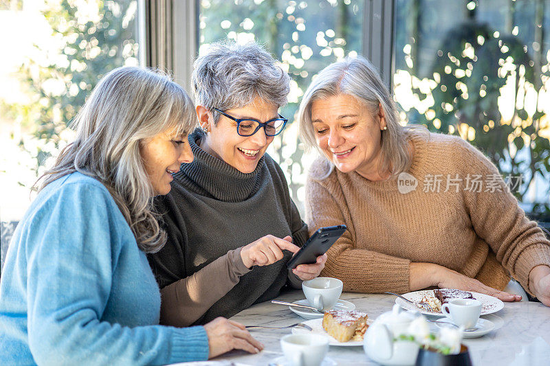 3个戴着白色帽子的微笑成熟女性朋友在咖啡馆吃早餐时正在使用智能手机。三位资深女性一起笑着看手机